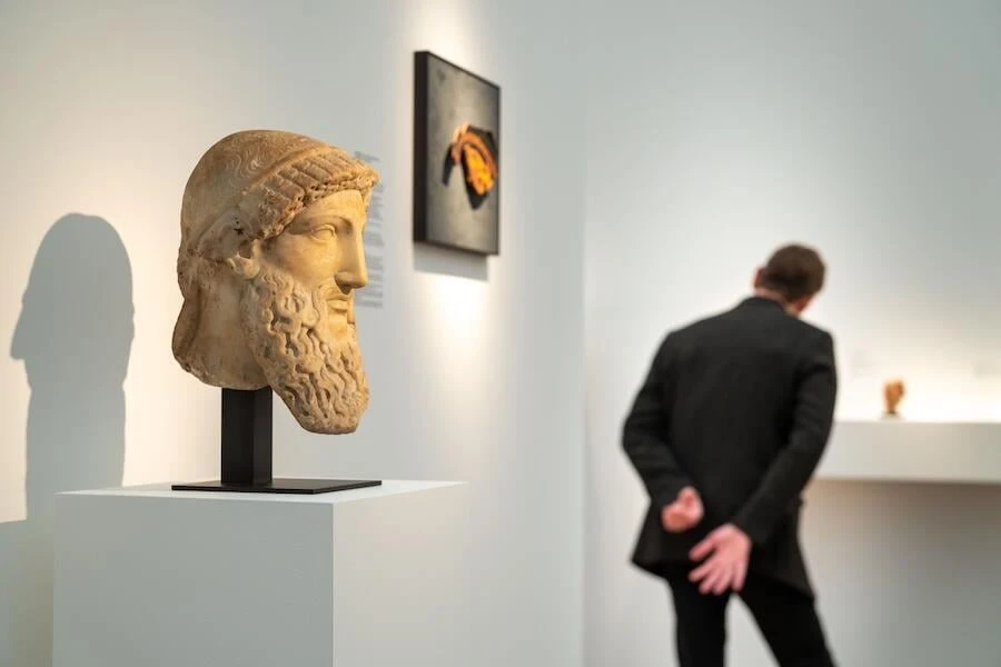 A classical marble head of a bearded man on a plinth, with a visitor looking at works further along the booth