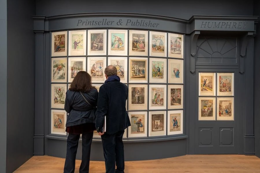 Two visitors studying a wall of framed eighteenth-century satirical prints in a recreated printseller’s shopfront